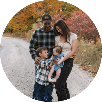 a family standing outdoors on a gravel path surrounded by vibrant autumn trees; two adults and two young children are present