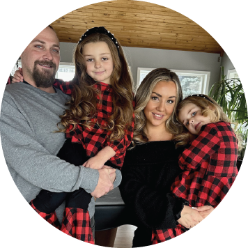 Two adults holding two children standing indoors, wearing coordinated red and black plaid tops, posed closely together in a bright room with windows and a wood ceiling.