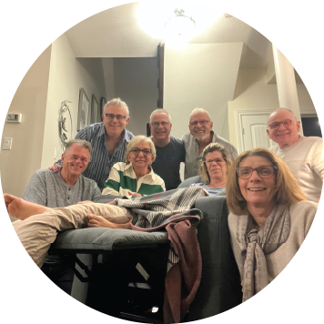 Smiling group of older adults gathered in a living room, surrounding a person resting on a couch