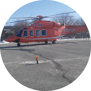 An orange Ornge helicopter parked on a snowy helipad. A person in a high-visibility jacket walks toward the aircraft. Trees and buildings are visible in the background under a clear blue sky.