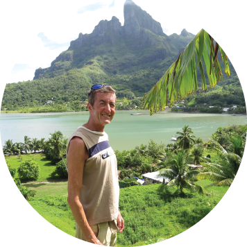 Person standing outdoors with a scenic view of a lush green landscape, palm trees, and a calm body of water with mountains in the background.