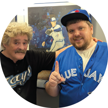 wo people wearing Toronto Blue Jays jerseys and caps pose in front of a photo of a baseball player, each holding up one finger.