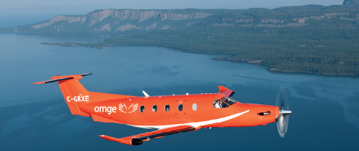 Un avion orange Ornge survolant un plan d'eau, avec des falaises boisées et un littoral en arrière-plan.