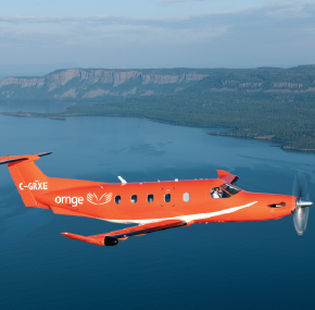 Un avion orange Ornge survolant un plan d'eau, avec des falaises boisées et un littoral en arrière-plan.