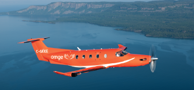 Un avion orange Ornge survolant un plan d'eau, avec des falaises boisées et un littoral en arrière-plan.