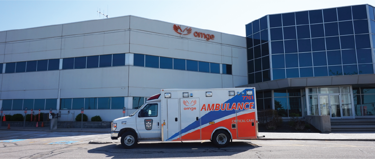Une ambulance Ornge stationnée devant un immeuble de bureaux avec de grandes fenêtres et le logo Ornge affiché bien en évidence sur la façade.