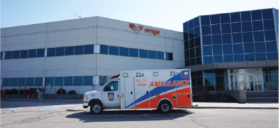Une ambulance Ornge stationnée devant un immeuble de bureaux avec de grandes fenêtres et le logo Ornge affiché bien en évidence sur la façade.