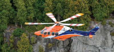 Orange and blue air ambulance helicopter flying past a rocky cliff with green forest in the background.