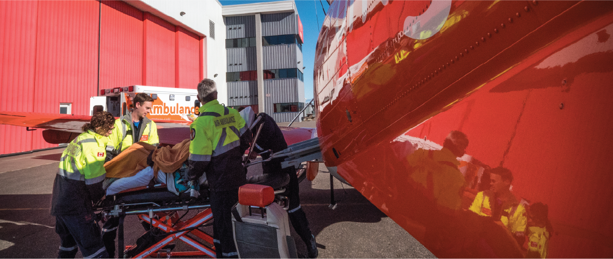 Des ambulanciers chargent un patient sur une civière dans une ambulance aérienne orange, à l'extérieur d'un hangar.