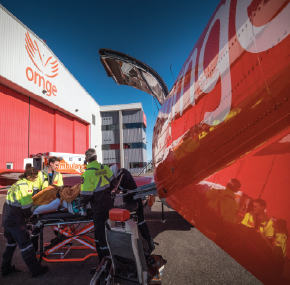 Des ambulanciers chargent un patient sur une civière dans une ambulance aérienne orange, à l'extérieur d'un hangar.