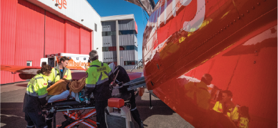 Des ambulanciers chargent un patient sur une civière dans une ambulance aérienne orange, à l'extérieur d'un hangar.