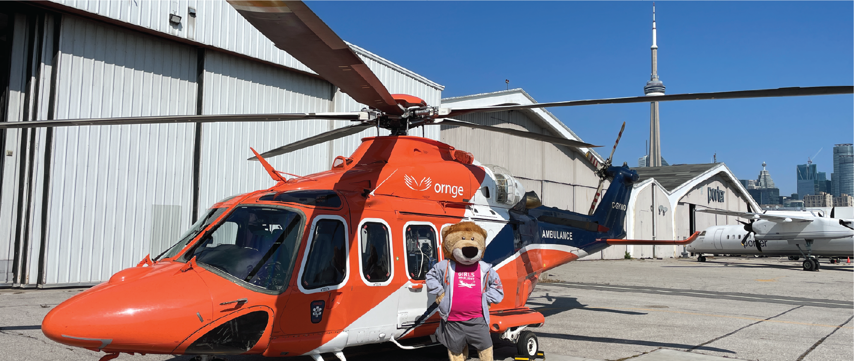 An orange helicopter parked on a tarmac with the Ornge Flyin Lion mascot standing in front, hangars and the Toronto skyline visible in the background.