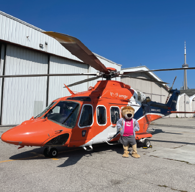 An orange helicopter parked on a tarmac with the Ornge Flyin Lion mascot standing in front, hangars and the Toronto skyline visible in the background.
