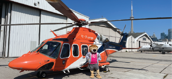 Un hélicoptère orange stationné sur le tarmac, avec la mascotte Orange Flyin Lion devant, des hangars et la silhouette de Toronto visibles en arrière-plan.