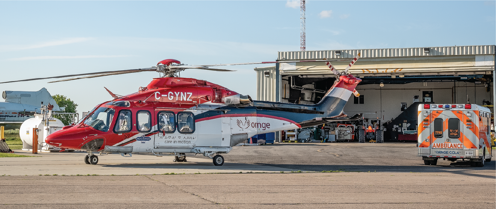 a red, white, and blue helicopter next to an ambulance parked outside of the Ornge hangar