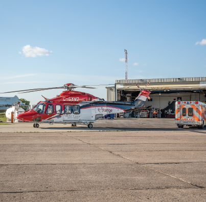 a red, white, and blue helicopter next to an ambulance parked outside of the Ornge hangar