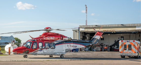a red, white, and blue helicopter next to an ambulance parked outside of the Ornge hangar