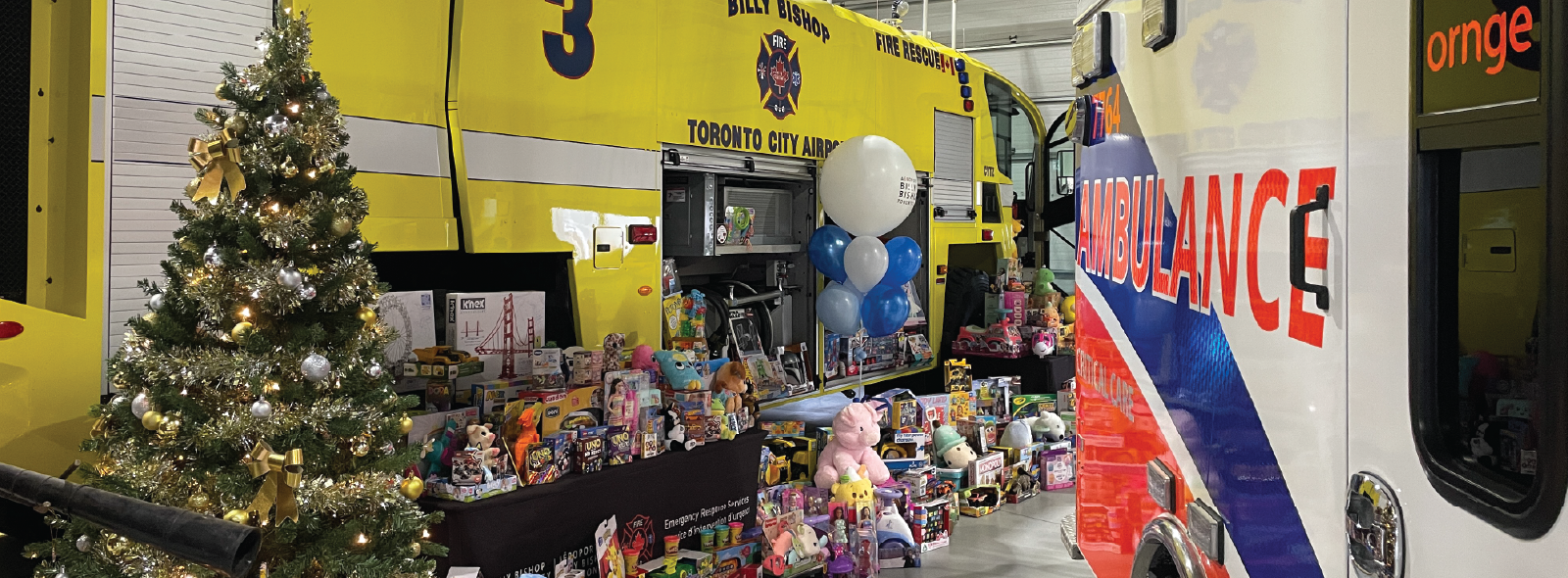 Christmas tree and toys displayed between a yellow fire truck and an Ornge ambulance inside a garage.