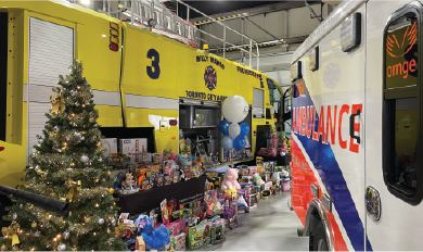 Christmas tree and toys displayed between a yellow fire truck and an Ornge ambulance inside a garage.