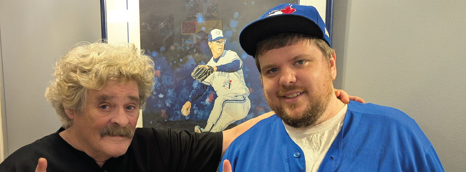 Two people wearing Toronto Blue Jays jerseys and caps pose in front of a photo of a baseball player, each holding up one finger.
