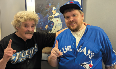 Two people wearing Toronto Blue Jays jerseys and caps pose in front of a photo of a baseball player, each holding up one finger.