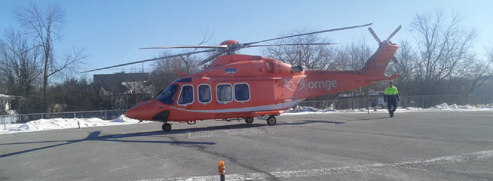 Un hélicoptère orange d'Ornge stationné sur une hélisurface enneigée. Une personne portant un gilet de haute visibilité s'approche de l'appareil. Des arbres et des bâtiments sont visibles en arrière-plan sous un ciel bleu clair.