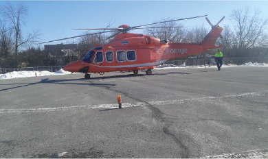 Un hélicoptère orange d'Ornge stationné sur une hélisurface enneigée. Une personne portant un gilet de haute visibilité s'approche de l'appareil. Des arbres et des bâtiments sont visibles en arrière-plan sous un ciel bleu clair.