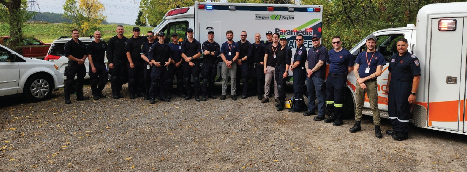 Group of uniformed paramedics standing in front of two ambulances on a gravel area surrounded by trees.