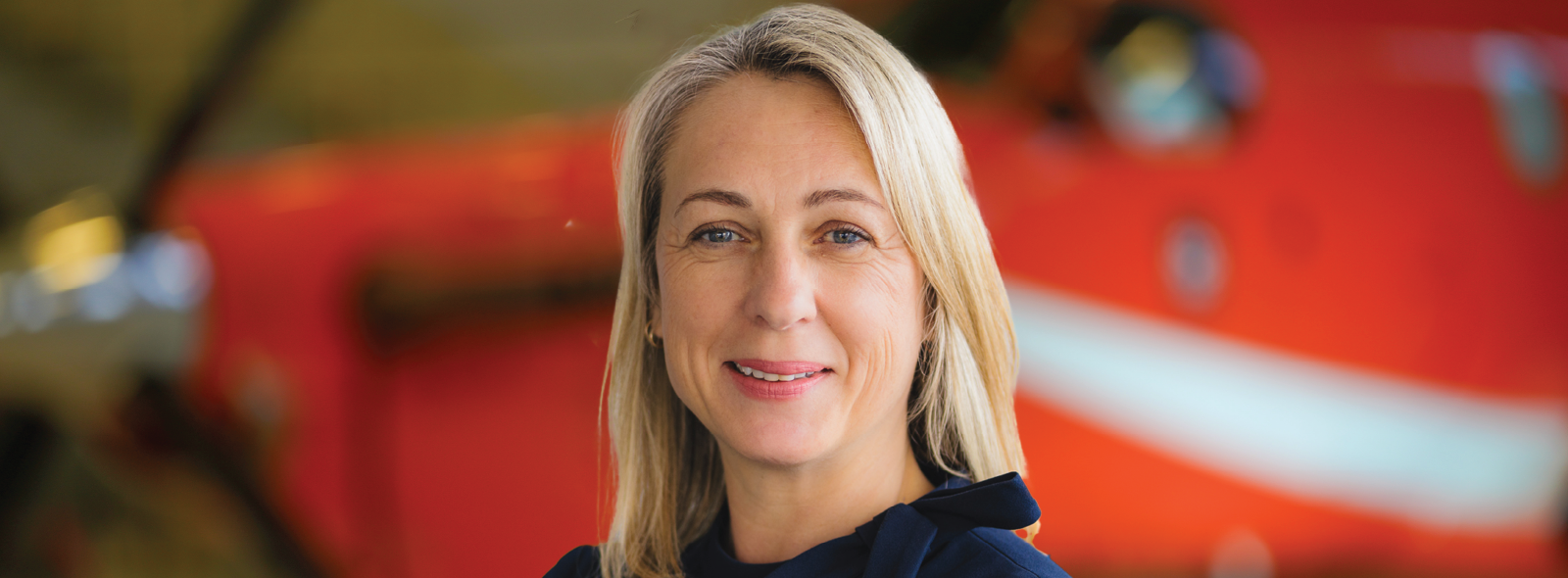 Woman with light-colored hair wearing a dark blue top, standing with arms crossed in front of a blurred orange aircraft