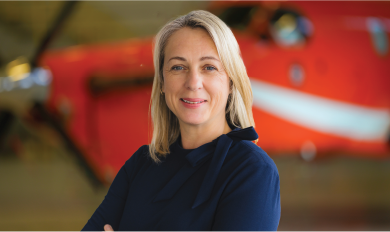 Woman with light-colored hair wearing a dark blue top, standing with arms crossed in front of a blurred orange aircraft