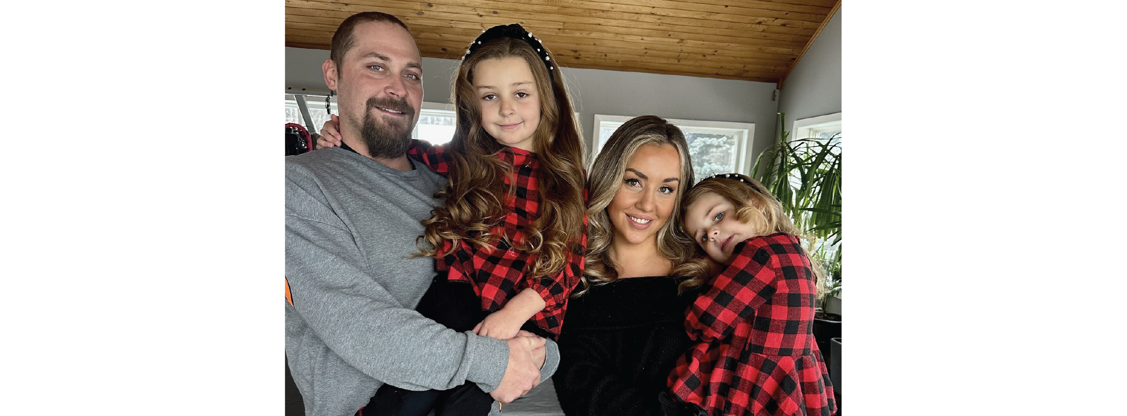 Two adults holding two children standing indoors, wearing coordinated red and black plaid tops, posed closely together in a bright room with windows and a wood ceiling.