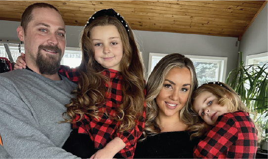 Deux adultes tenant deux enfants, debout à l'intérieur, vêtus de hauts à carreaux rouges et noirs assortis, posent côte à côte dans une pièce lumineuse avec des fenêtres et un plafond en bois.