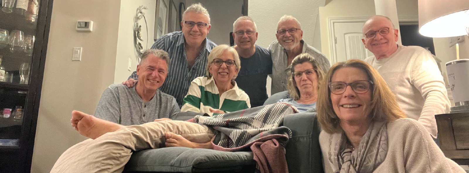Smiling group of older adults gathered in a living room, surrounding a person resting on a couch.