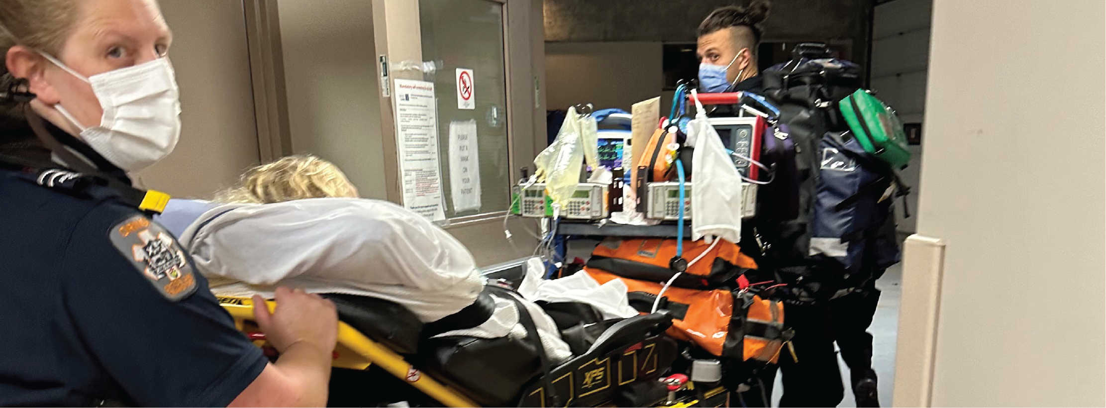 Paramedics transport a patient on a stretcher through a hospital hallway with medical equipment attached.