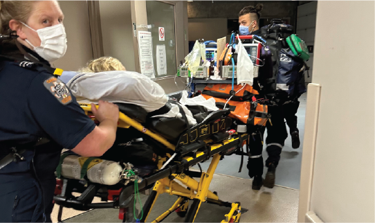 Paramedics transport a patient on a stretcher through a hospital hallway with medical equipment attached.