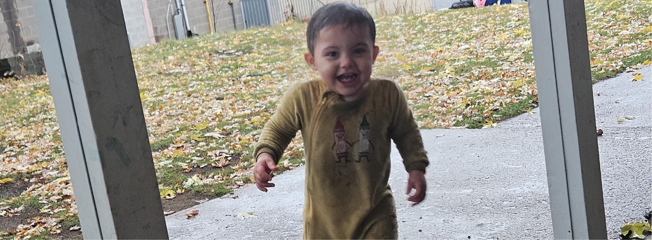 A toddler stands on a wet patio with fallen leaves scattered across the yard in the background.