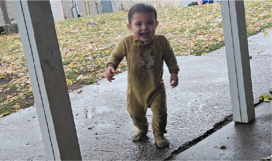 A toddler stands on a wet patio with fallen leaves scattered across the yard in the background.