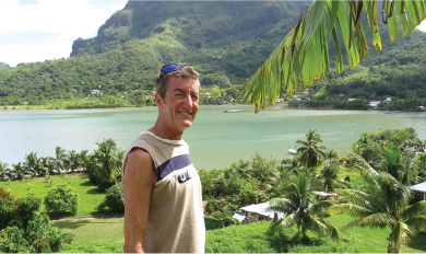 Person standing outdoors with a scenic view of a lush green landscape, palm trees, and a calm body of water with mountains in the background.