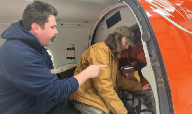 Deux personnes sont assises à la porte ouverte d'un avion orange, l'une d'elles désignant du doigt un équipement à l'intérieur de la cabine.