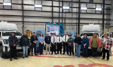 Group of people standing in a hangar between two medical transport vehicles with a blue emergency response truck in the background.