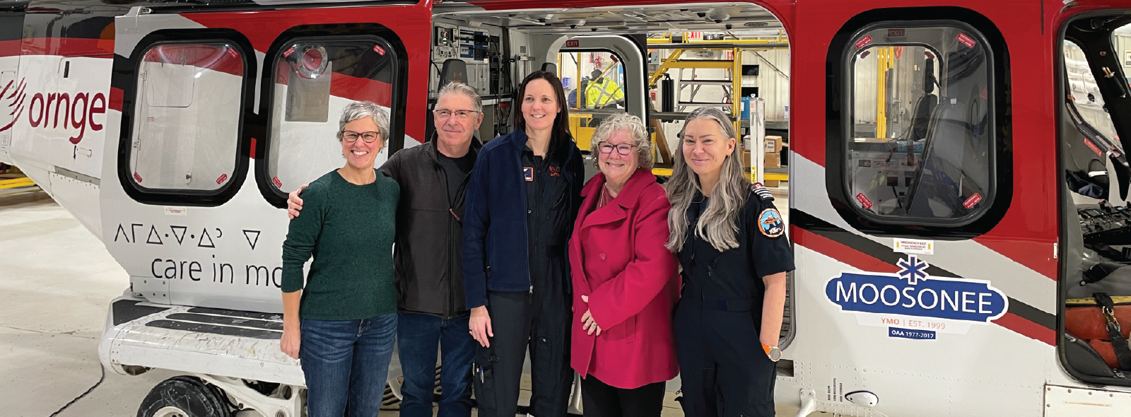 Five people standing in front of a red and white Ornge helicopter inside a hangar