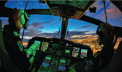 View from inside a helicopter cockpit with two crew members and illuminated flight controls as the aircraft flies over a city