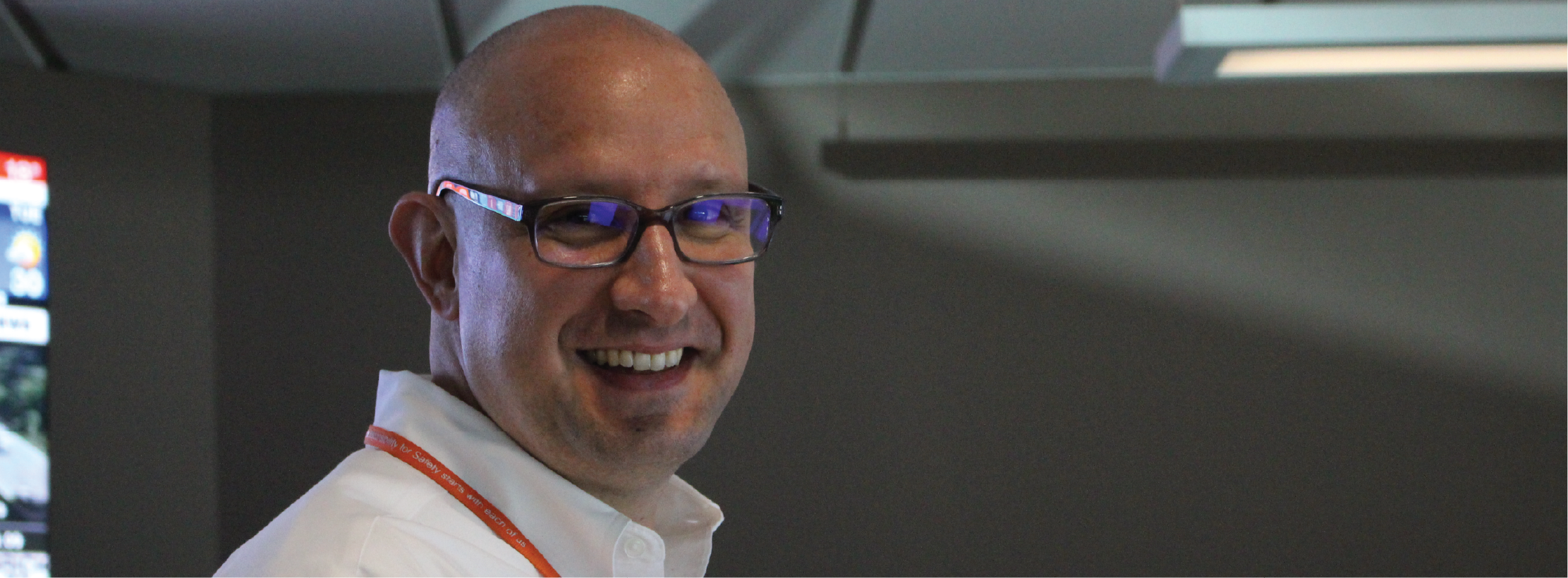 a man with glasses and a white shirt standing in front of a computer screen and smiling at the camera