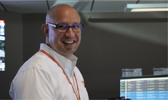 a man with glasses and a white shirt standing in front of a computer screen and smiling at the camera