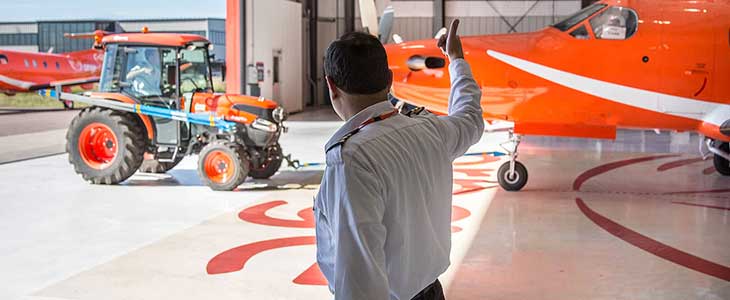 Man giving thumbs up in foreground, in background a tractor with a fixed wing Ornge plane