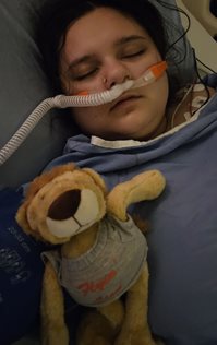 A patient lying in a hospital bed with medical tubing attached, holding a small stuffed lion toy.