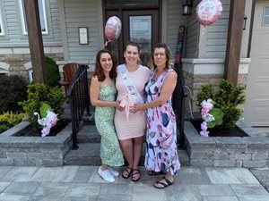 Three women stand together on a front porch, smiling and holding a pink balloon and sash at a baby shower