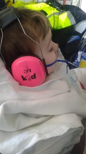 A close-up of a child resting with a clear oxygen mask and pink earmuffs labeled 'P1'. The child is wrapped in a white blanket, with medical tubing visible. A high-visibility jacket with yellow and reflective stripes is partially seen in the background.
