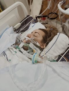 Patient lying in a hospital bed with breathing tubes and medical equipment visible around the head and chest.